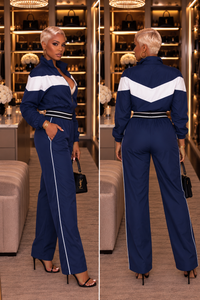 A woman modeling a navy blue and white athletic jumpsuit, shown in front and back views. The jumpsuit features a high collar, a white V-shaped chest stripe, and white piping running down the sides of the wide-leg pants.